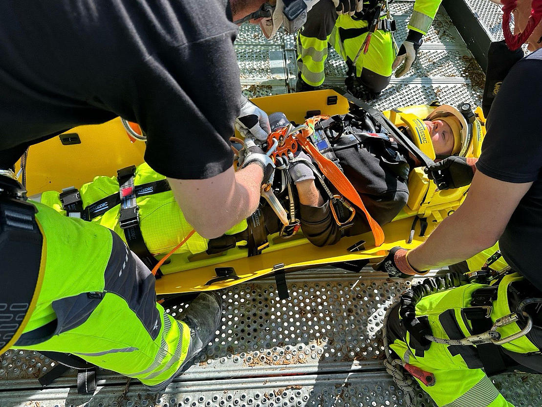Sicherung in der Rettungstrage – praktische Rettungsschulung Schulungsteilnehmer sichern eine Person mit Gurten in einer Rettungstrage auf einer Gerüstplattform.